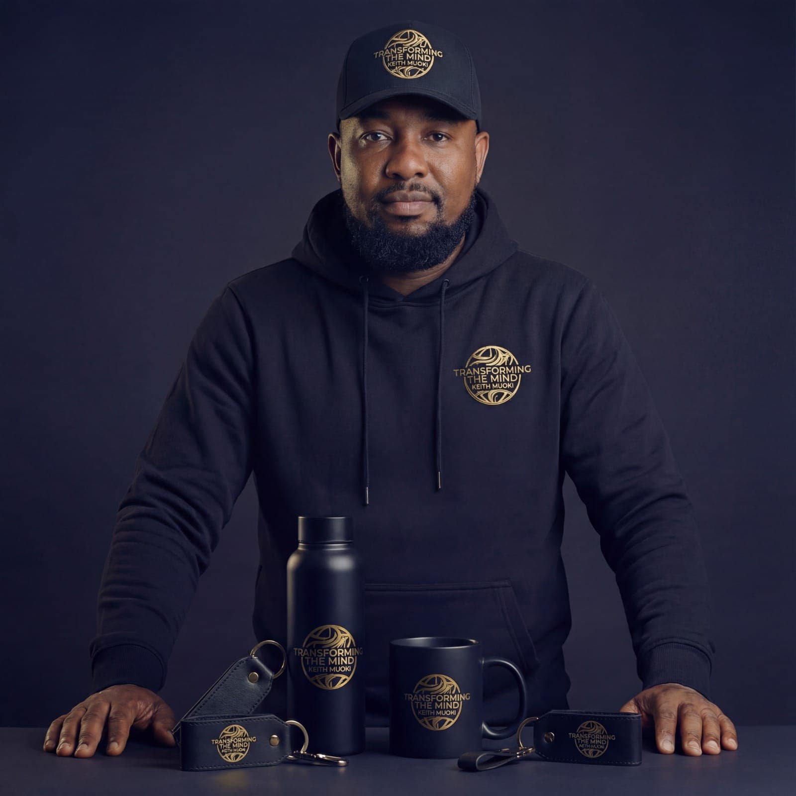 Studio portrait of Keith Muoki in a Transforming the Mind hoodie and cap, standing behind a table of matching branded merchandise: a water bottle, a mug and leather keyrings
