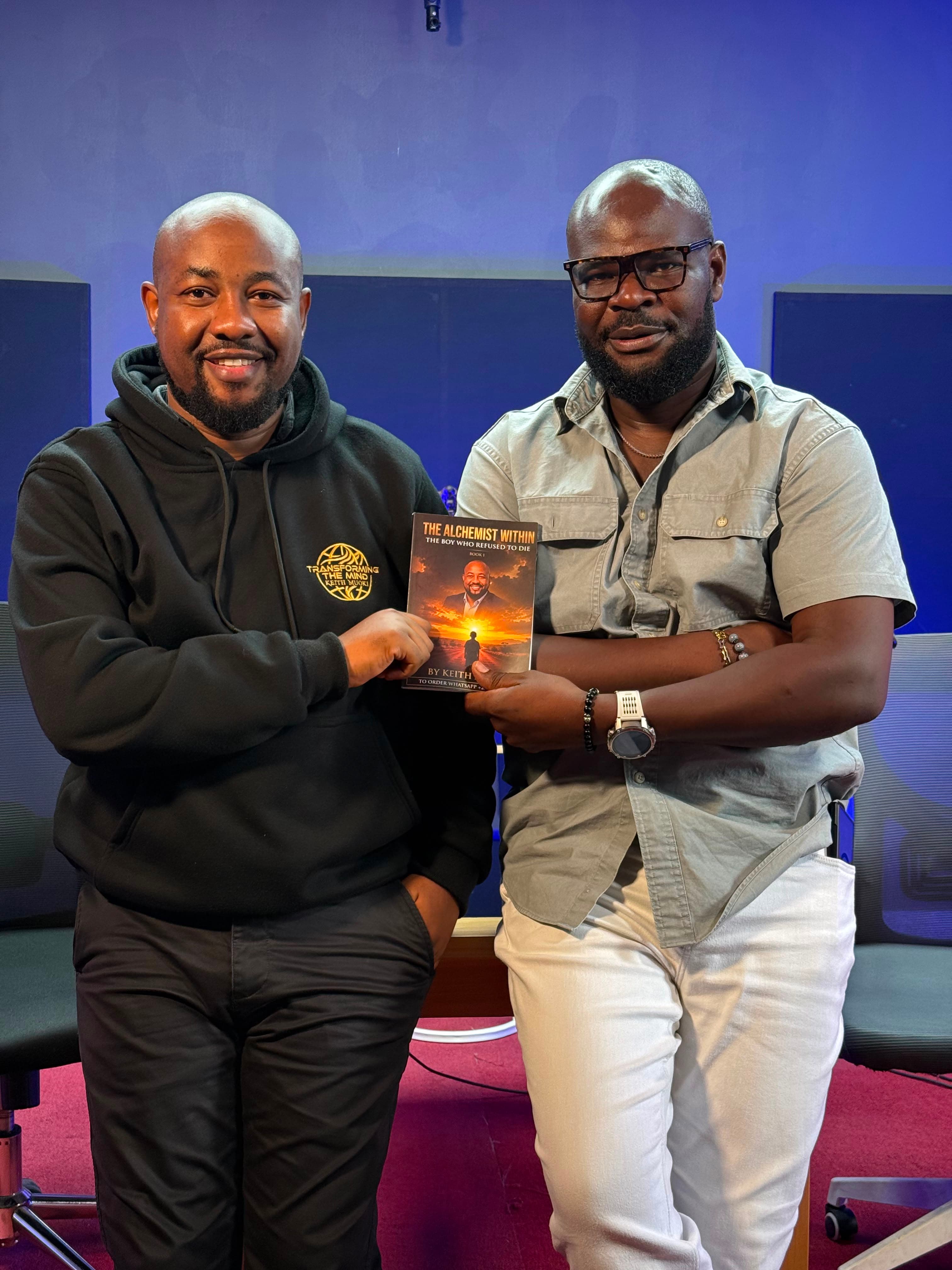 Keith Muoki, in a Transforming the Mind hoodie, holding a copy of his book The Alchemist Within: The Boy Who Refused To Die alongside a fellow guest in a studio