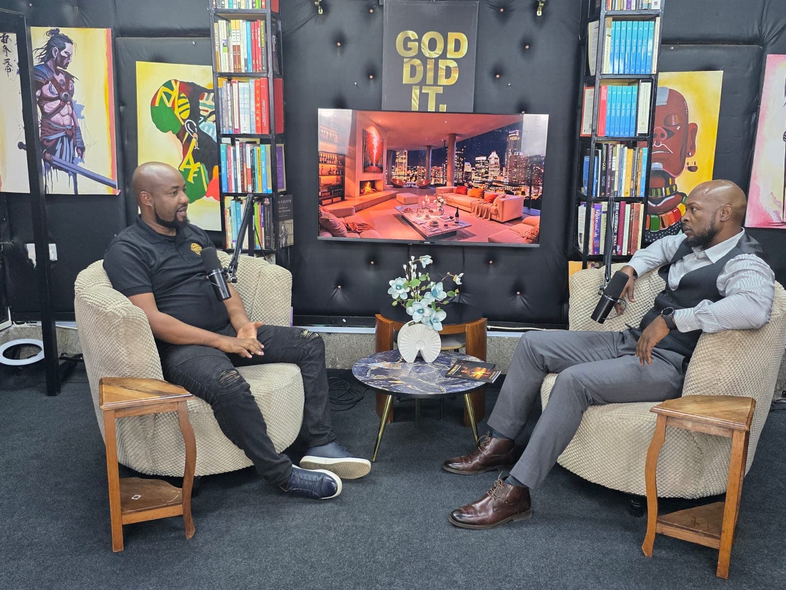 Keith Muoki and his interviewer seated in armchairs facing each other on a podcast set, boom microphones between them and bookshelves and painted artwork behind