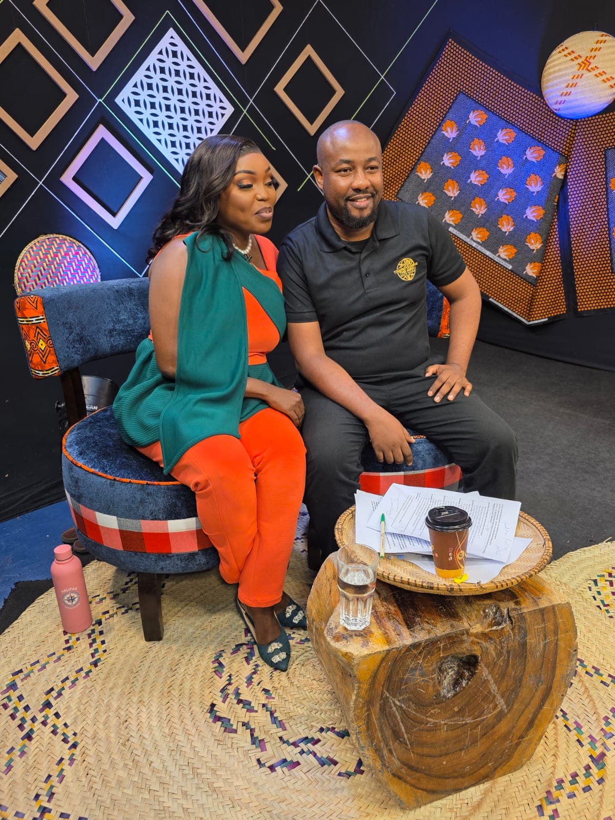 Keith Muoki seated beside a presenter on a television set decorated with African-print panels and woven baskets, with the interviewer’s notes on a wooden side table