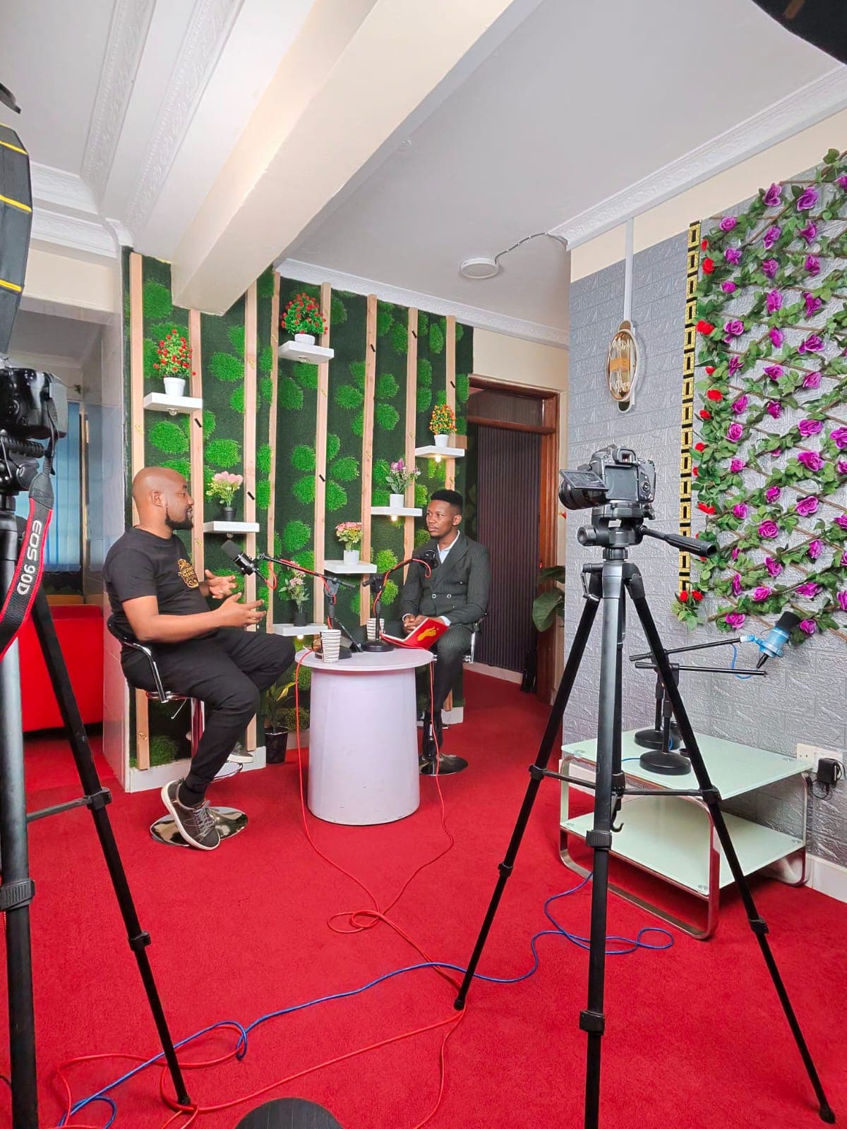 A podcast in production: two cameras on tripods and a lighting stand trained on Keith Muoki and his interviewer, who face each other across a small table of microphones